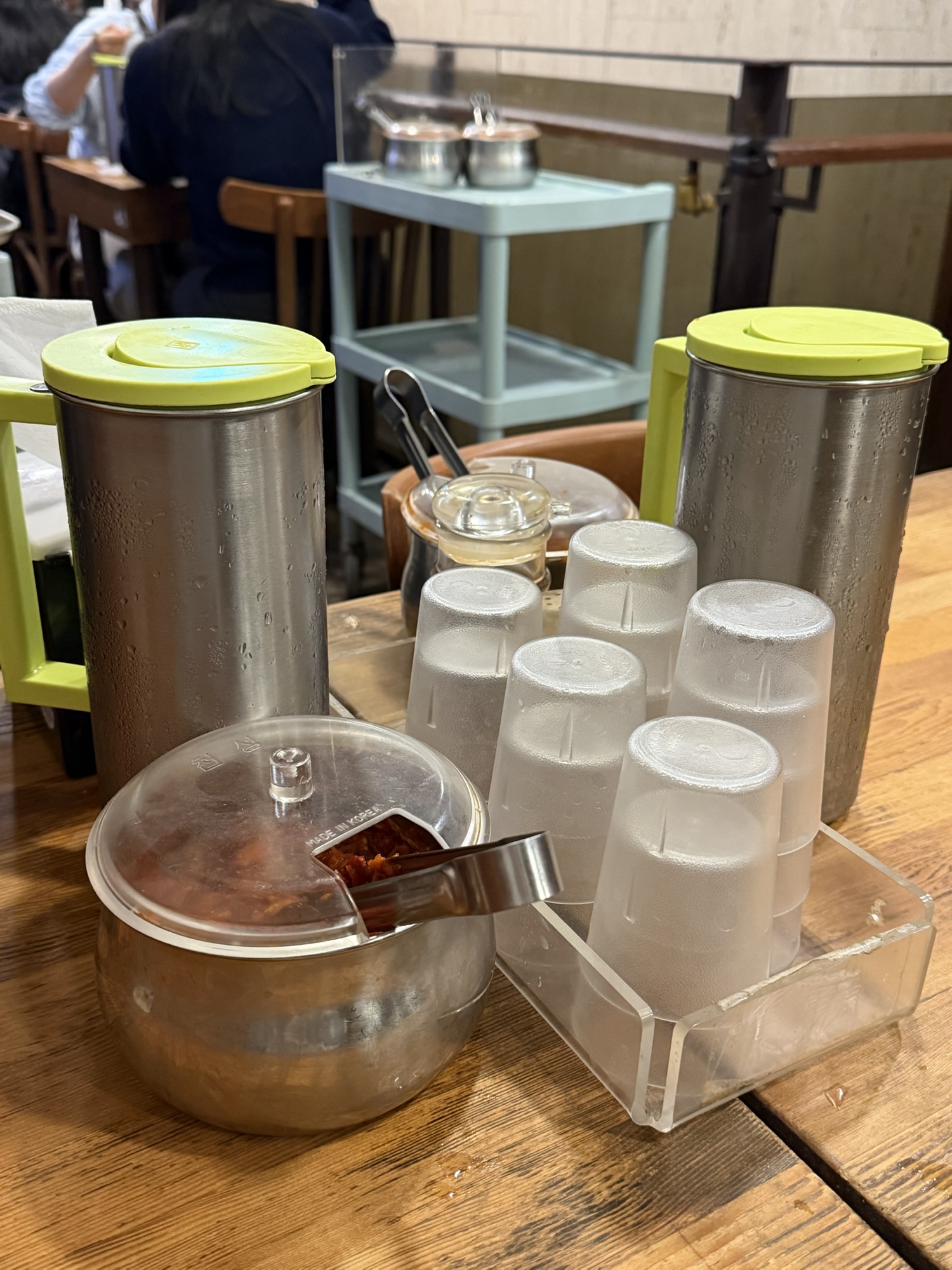 Tabletop water pitchers, plastic cups, and a glass jar of garlic kimchi at Myeongdong Kyoja