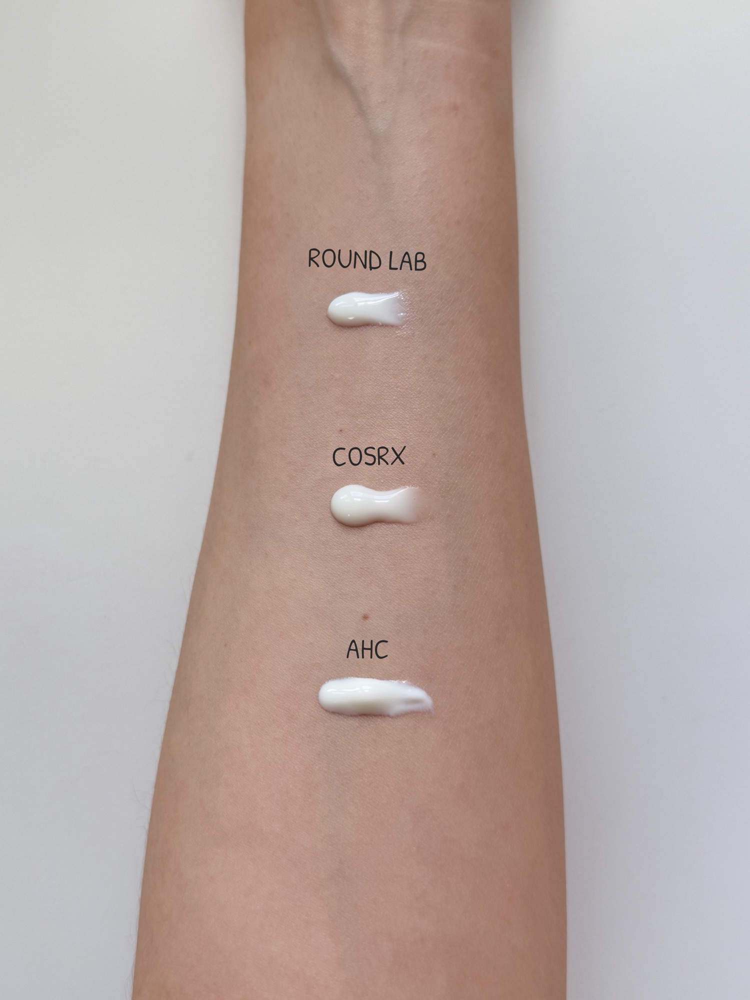 Three sunscreens compared side by side on the back of hand — ROUND LAB, COSRX, AHC texture comparison before spreading