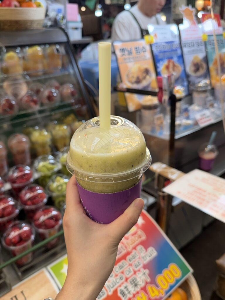 Cup of pale green kiwi-and-pineapple fresh juice from a Gwangjang Market stall, held in front of the fruit display