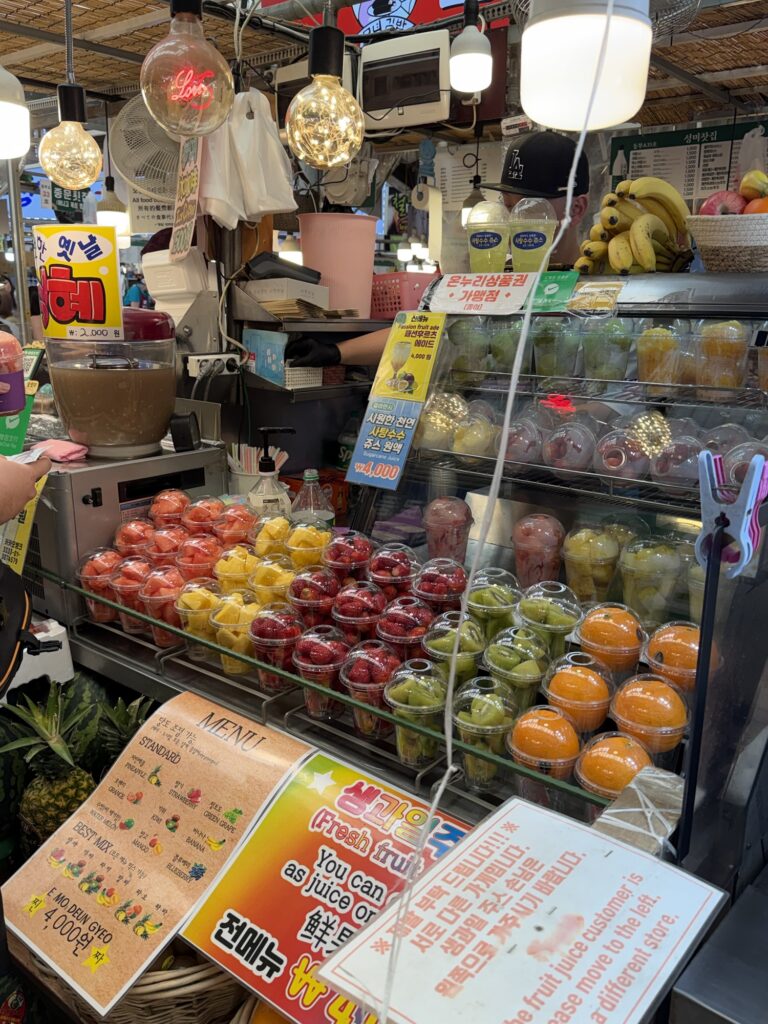 Fresh fruit juice stall at Gwangjang Market with cups of pre-cut fruit lined up — strawberries, kiwi, oranges, pineapple — and a ₩4,000 sign