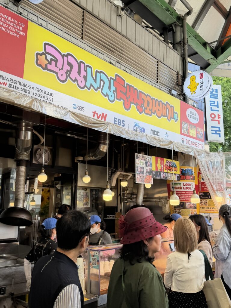 Gwangjang Market Chapssal Kkwabaegi storefront with the famous pink and yellow signage and broadcast network logos (KBS, tvN, EBS, MBC) above the queue