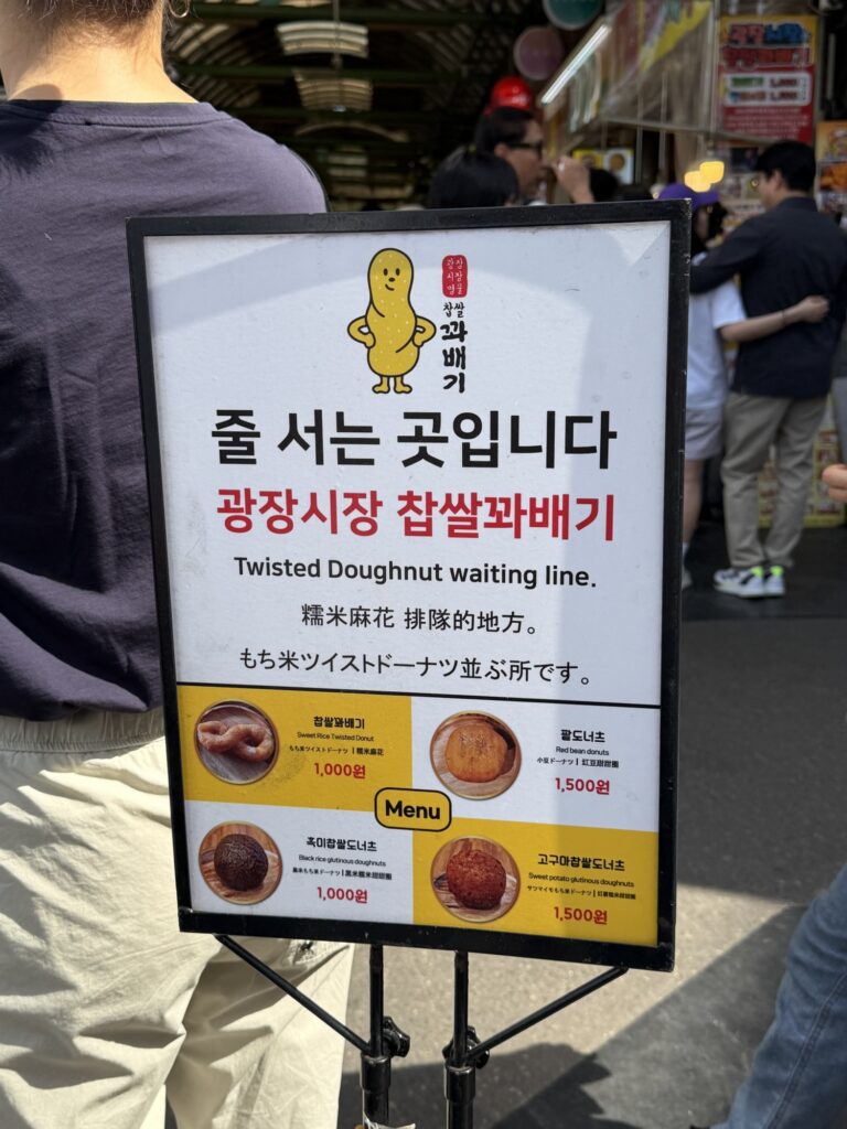 Gwangjang Market Chapssal Kkwabaegi queue sign with English, Chinese, and Japanese translations and a menu showing twisted donut ₩1,000, red bean donut ₩1,500, black rice donut ₩1,000, sweet potato donut ₩1,500