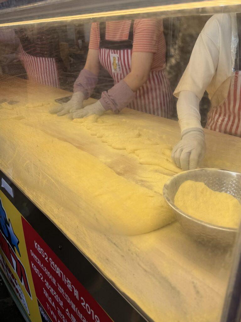 Two workers in striped aprons shaping a long block of yellow glutinous-rice dough at Gwangjang Market Chapssal Kkwabaegi — production-line style