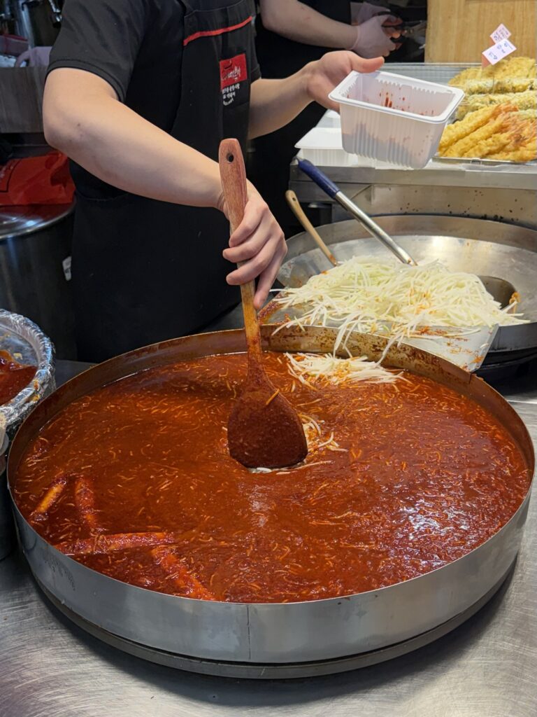 Giant round pan of red Busan-style tteokbokki at Gwangjang Market topped with shredded white radish, being stirred with a wooden paddle