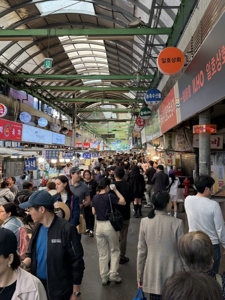 Gwangjang Market main aisle on a Sunday afternoon — arched roof, neon signs, dense crowd of shoppers and snack hunters